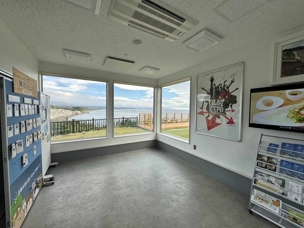 乙部町・滝瀬海岸シラフラ展望公園の休憩所内部。
大きな窓からシラフラ海岸と日本海の絶景が一望できる。
壁にはGLAY EXPOのポスターが掲示され、
左側には乙部町の観光カードや観光パンフレットが並ぶ。
モニターには地元の海産物（ウニなど）の映像が映っている。
