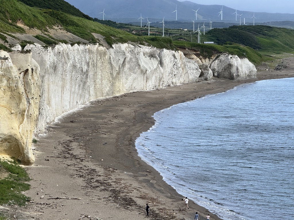 北海道乙部町・滝瀬海岸シラフラの白い断崖絶壁が
約500mにわたって続く絶景。
青い空と緑の丘、砂浜と日本海のコントラストが美しく、
遠景には風力発電の風車が並ぶ。
数名の観光客が砂浜を歩いており、スケール感が伝わる一枚。
