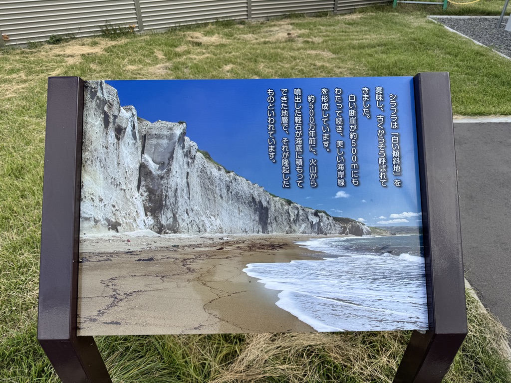 乙部町・シラフラの現地説明板。
アイヌ語で「白い傾斜地」を意味するシラフラの由来と、
約500万年前の火山噴火による地層形成の歴史が
日本語で記されている。
パネルには青空を背景にした白い断崖の写真が使用されている。
